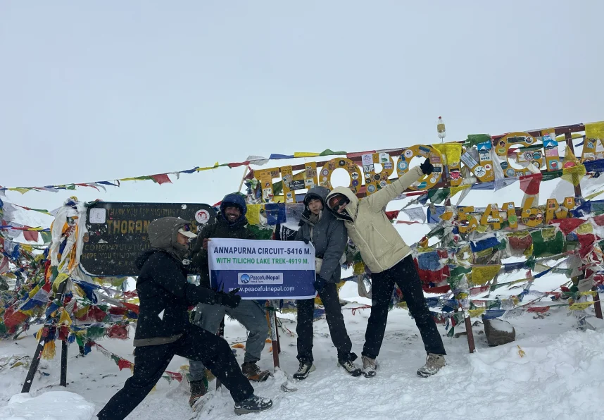 Trekkers posing at Thorong La Pass on the Annapurna Circuit during snowy weather with prayer flags and signboard