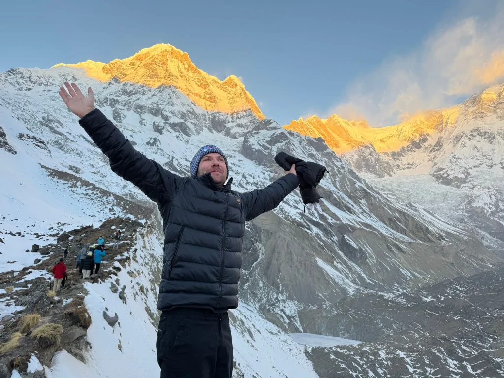 Man celebrating at Annapurna Base Camp with golden sunrise light on Annapurna peaks and snowy Himalayan landscape in Nepal.