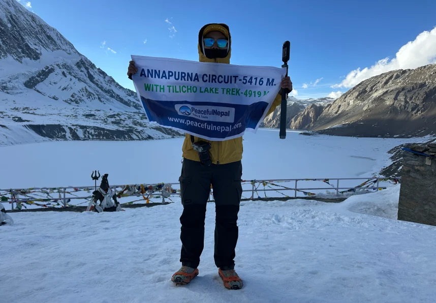 Trekker holding banner at Annapurna Circuit near Tilicho Lake with snowy mountains and frozen lake in the background