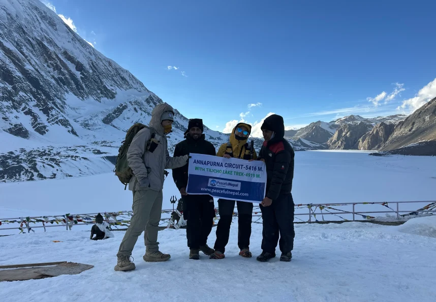 Group of trekkers posing with a banner at Tilicho Lake during the Annapurna Circuit trek, with snow-covered mountains and a clear blue sky.