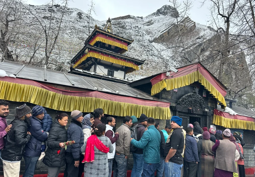 Pilgrims standing in a queue outside Muktinath Temple in Nepal, surrounded by snow-covered mountains and leafless trees, with traditional pagoda-style architecture decorated in red and yellow fabric.
