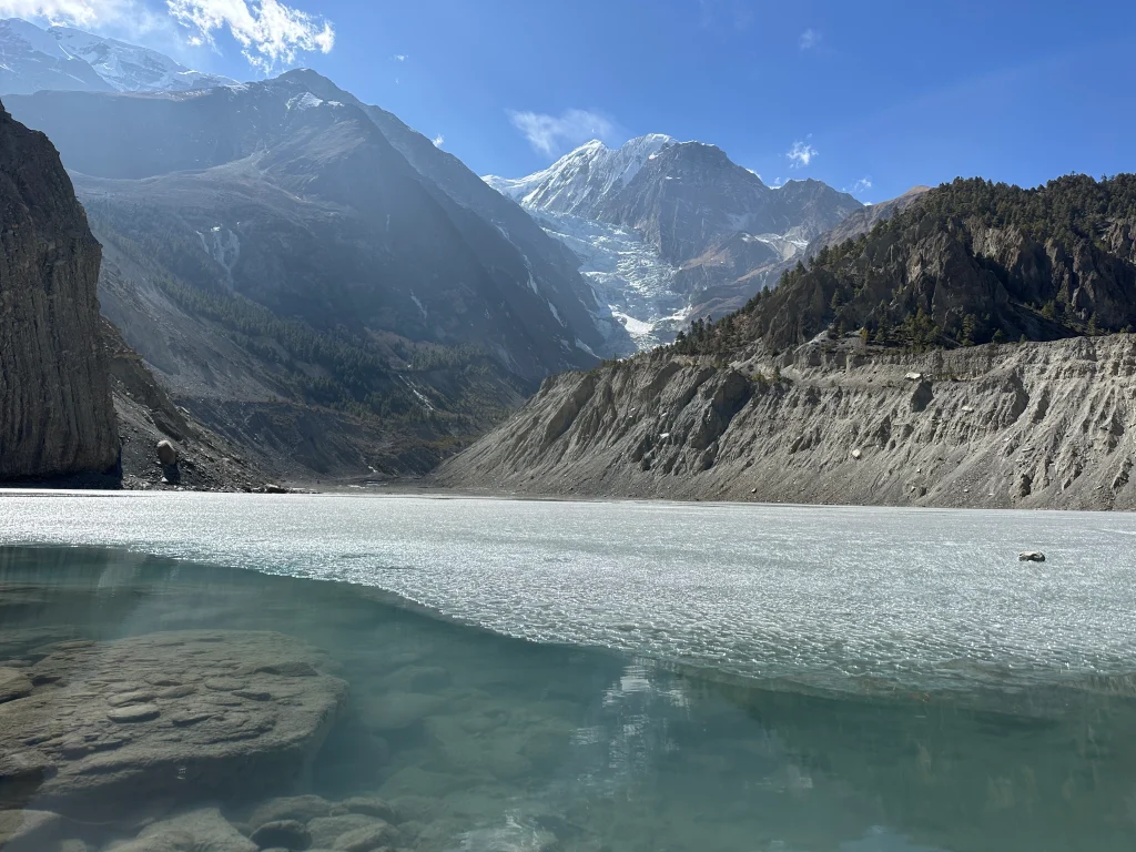 Frozen Gangapurna Lake in Manang, Nepal, with crystal-clear water, dramatic rocky cliffs.