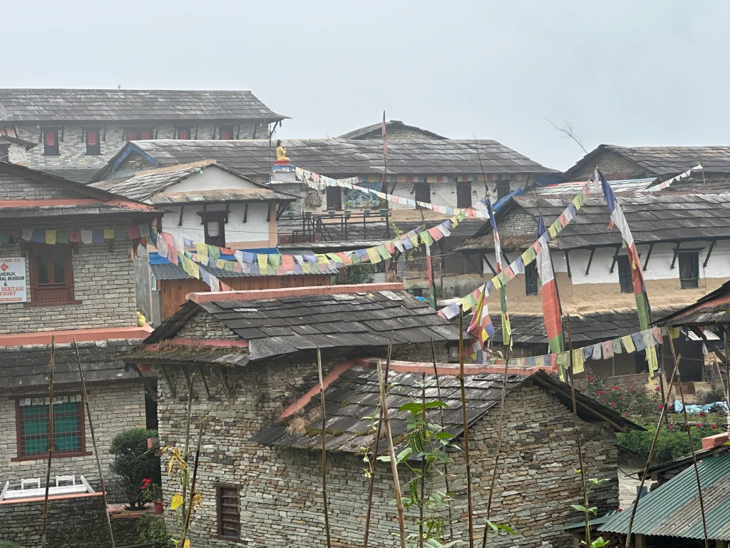 Colorful prayer flags over traditional stone houses in Ghandruk Gurung village with misty mountain backdrop.