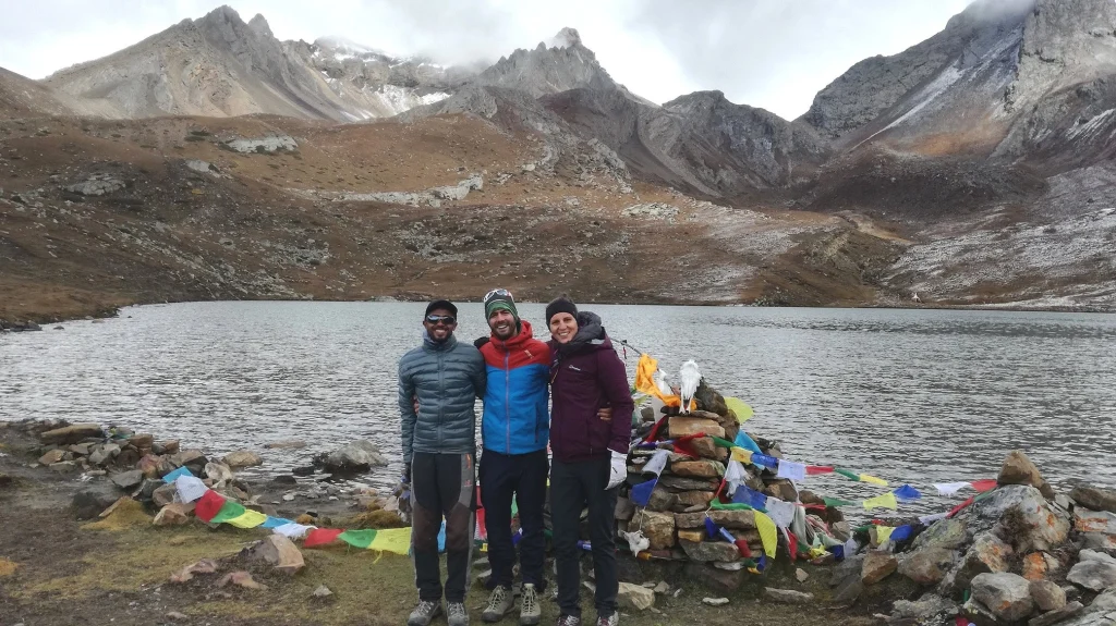Three trekkers standing beside a high-altitude ice lake with colorful prayer flags and rugged Himalayan mountains in the background.