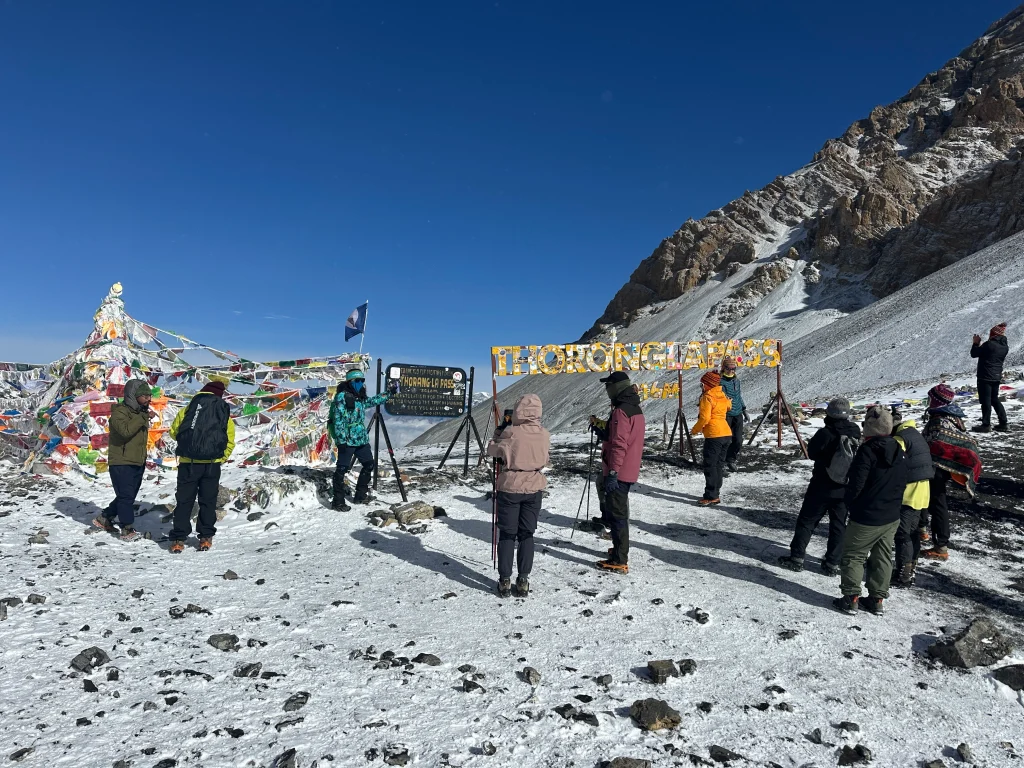 Trekkers at Thorong La Pass (5,416m) with prayer flags and snowy Himalayan landscape in Nepal.