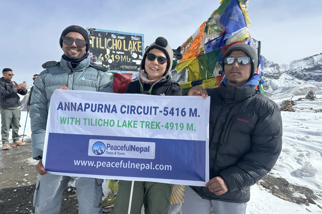 Three trekkers holding a Peaceful Nepal Treks & Tours banner at Tilicho Lake (4,919 m) during the Annapurna Circuit Trek,