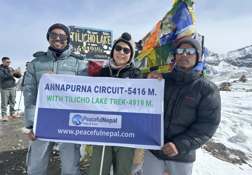 Three trekkers holding a Peaceful Nepal Treks & Tours banner at Tilicho Lake (4,919 m) during the Annapurna Circuit Trek,